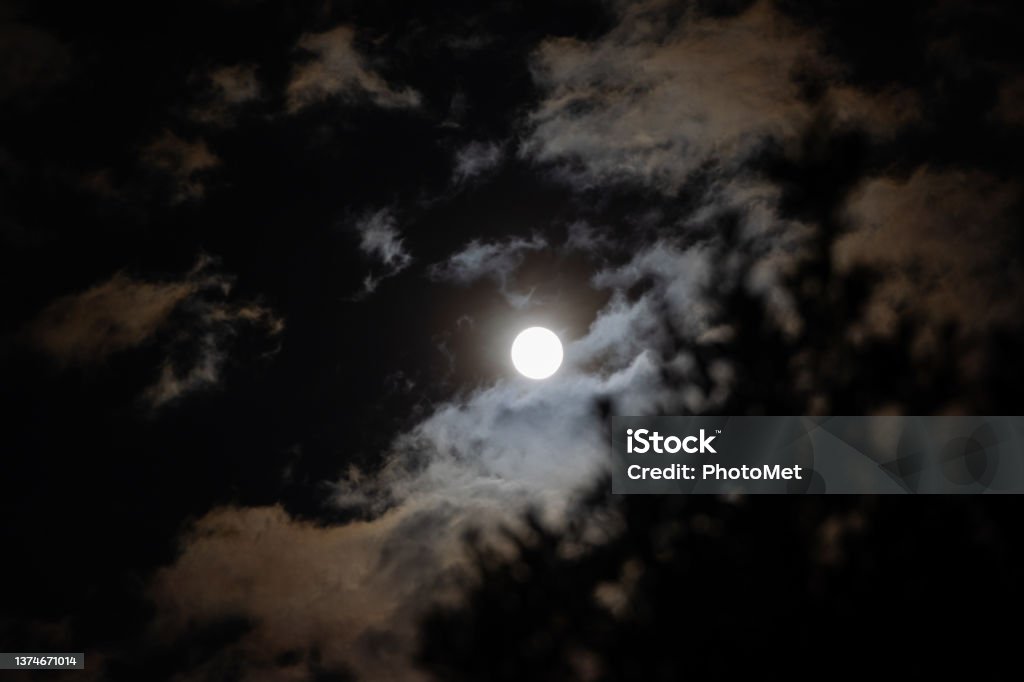 full white moon seen from behind little clouds at night in the dark sky close up view of the full white moon seen from behind little clouds at night in the dark sky Night Stock Photo