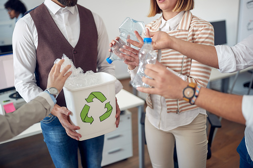 A group of employees is collecting plastic bottles in the recycling bin in a pleasant atmosphere in the office. Employees, job, office