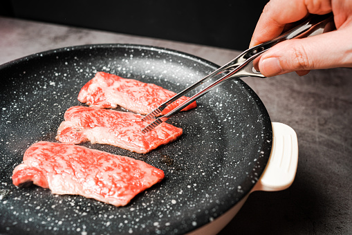 Premium Rare Slices sirloin Wagyu A5 beef with high-marbling texture pick up and grill on marble coated non-stick pan by bbq tongs with hand on black background. Served for Yakiniku.