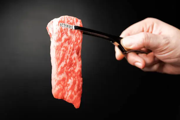 Premium Rare Slices sirloin Wagyu A5 beef with high-marbling texture pick up by bbq tongs with hand on black background. Served for Yakiniku, Sukiyaki and Shabu. Image with copy space.