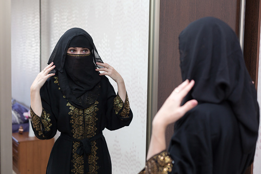 A Muslim woman dresses up in front mirror in her apartment, adjusting her burqa.