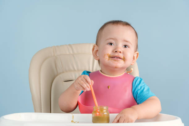 istock Toddler eats broccoli vegetable puree with an orange plastic spoon while sitting on a feeding chair 1374785319
