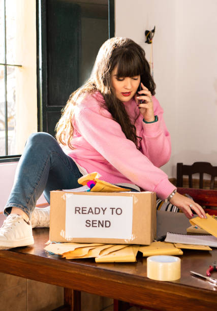 Female worker sitting on table and having conversation on cellphone and preparing orders in envelopes for sending in boxes in handicraft workshop