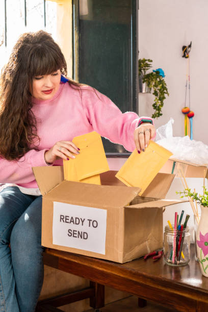 istock Female maker sitting on table and preparing paper envelopes with orders for sending in carton box in handicraft workshop 1374810327