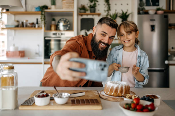 Father and daughter taking a selfie while decorating a cake in the kitchen stock photo
