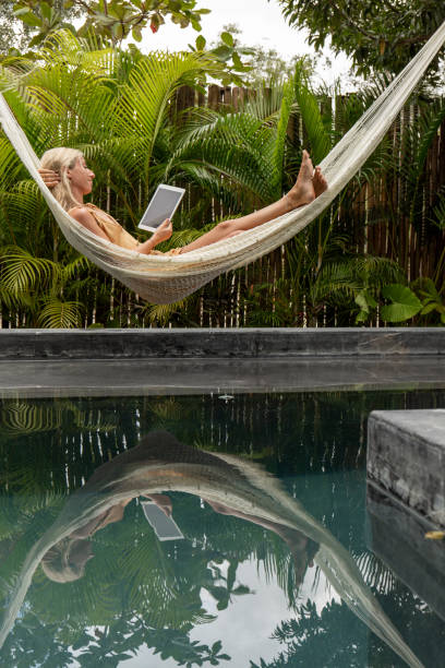 Woman relaxing in an hammock by the pool with a digital tablet 30's woman relaxing on hammock by the pool in a tropical garden using a digital pad reading, purchasing, booking. digital nomad stock pictures, royalty-free photos & images