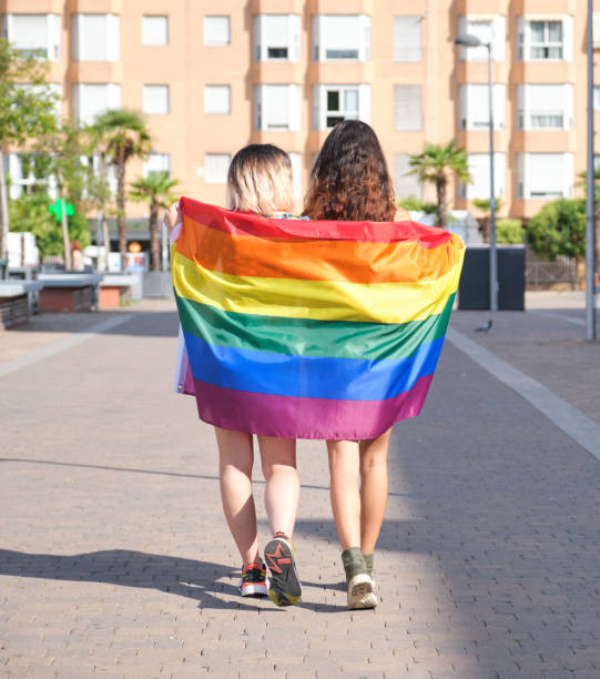 jovem lésbica andando enrolada em uma bandeira do arco-íris para trás. - lesbian love sexual issues two people - fotografias e filmes do acervo