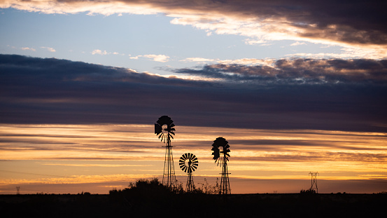 Three Wind-powered pumps Windmills Windpomp at sunset with Electricity Cables silhouette.  A windpumps are used on farms using the wind to pump up water from the water tables below the ground.  It is being used a lot in the Karoo and Kalahari region of Southern Africa.