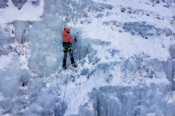 An ice climber in a yellow jacket and blue helmet looking downward, perhaps looking for one of his tools.  Against the snowy wild background, the image shows the quiet and waiting times that are part of climbing.