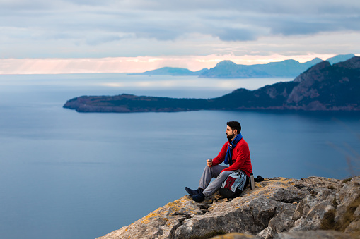 Hiker with red sweatshirt sitting on a rock in front of the beautiful north coast of Mallorca during the sunrise drinking hot tea from a thermos flask. Color editing. Part of a series.