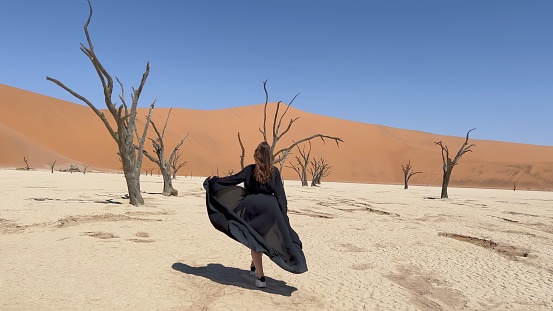 A young woman in a long black dress walks through the desert. Dried up ancient lake with dead trees in Africa. Mystical landscape, witch. Dead vlei, Namibia, Sossusvlei. Strong wind, girl waving gown.