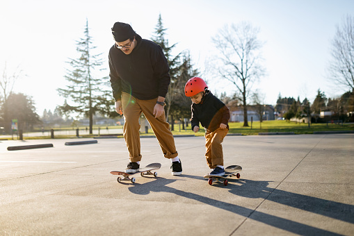 A Hispanic dad rides a skateboard with his 5 year old child, enjoying the time together.