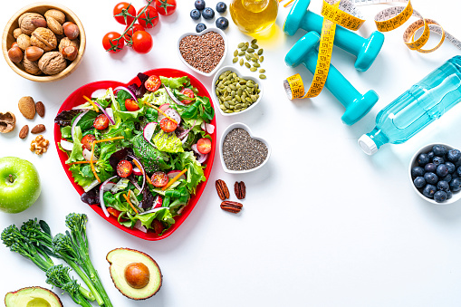 Overhead view of a red heart shape plate with healthy salad shot on white background. Dumbbells and a tape measure are at the top right. Useful copy space available at the right. Multi colored fresh fruits, vegetables, seeds and nuts are around the salad plate. High resolution 42Mp studio digital capture taken with SONY A7rII and Zeiss Batis 40mm F2.0 CF lens