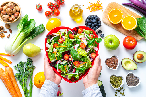 Overhead view of female hands holding a red heart shape plate with healthy salad shot on white background. Multi colored fresh fruits, vegetables, seeds and nuts are out of focus at background. High resolution 42Mp studio digital capture taken with SONY A7rII and Zeiss Batis 40mm F2.0 CF lens