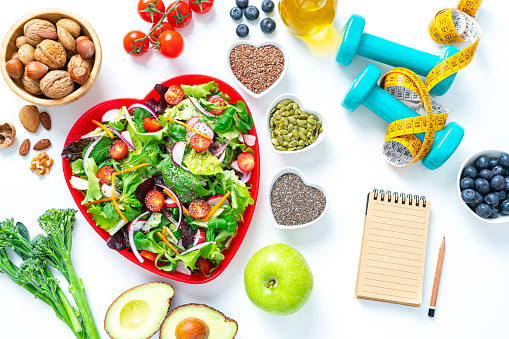 Overhead view of a red heart shape plate with healthy salad shot on white background. Dumbbells and a tape measure are at the top right. A blank note pad with useful copy space is included in the composition. Multi colored fresh fruits, vegetables, seeds and nuts are around the salad plate. High resolution 42Mp studio digital capture taken with SONY A7rII and Zeiss Batis 40mm F2.0 CF lens