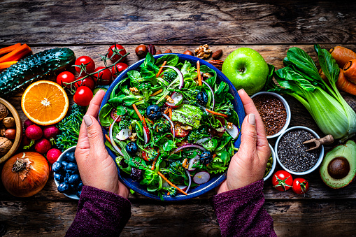 Overhead view of female hands holding a blue plate with healthy salad shot on rustic wooden table. Multi colored fresh fruits, vegetables, seeds and nuts are at background. High resolution 42Mp studio digital capture taken with SONY A7rII and Zeiss Batis 40mm F2.0 CF lens
