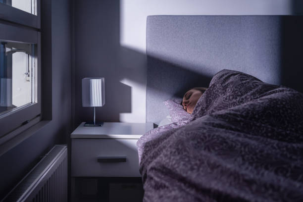 Sleeping woman in bed at night in dark bedroom. Person resting. Asleep under cover and blanket. Cold room at home. Tired lady in peaceful dream. Blue moonlight from window. stock photo