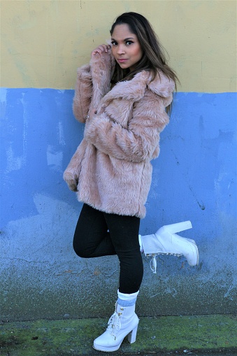 A woman posing against a yellow and blue grungy wall. She is wearing a fur coat, black pants, whit socks and boots.