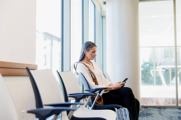 Woman uses smart phone while sitting in waiting room The mid adult woman uses her smart phone as she sits in the hospital waiting room. waiting room stock pictures, royalty-free photos & images