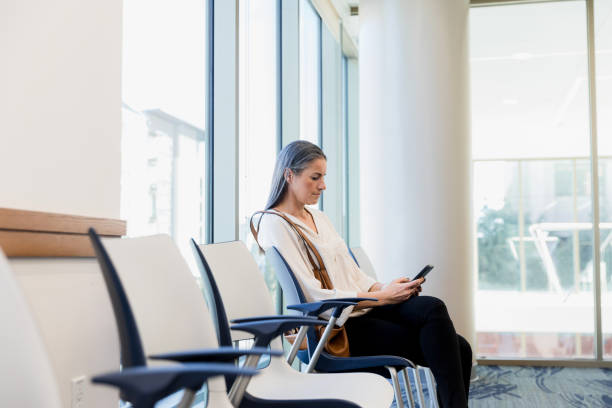 Woman uses smart phone while sitting in waiting room The mid adult woman uses her smart phone as she sits in the hospital waiting room. medical office lobby stock pictures, royalty-free photos & images