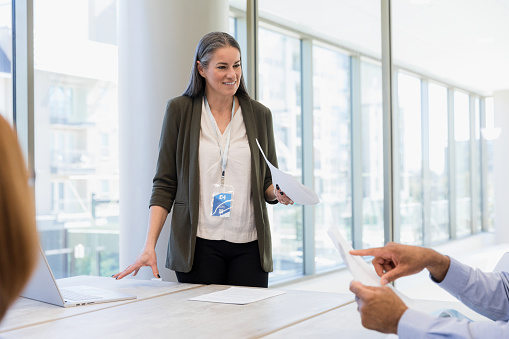 The mid adult woman leading the team meeting listens to the question from the unrecognizable man as he points to the document in his hand.