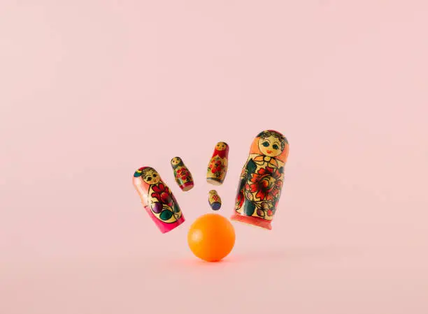 Russian dolls known as matryoshka or babushka as the pins flying from the strike of the orange bowling ball on a pink background. Minimal concept.