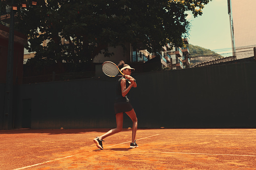 A sportswoman holding a tennis racket over her shoulder, with her feet flexed and looking straight ahead. An African woman practicing tennis attacks alone outdoors. The lady exercising on a game court
