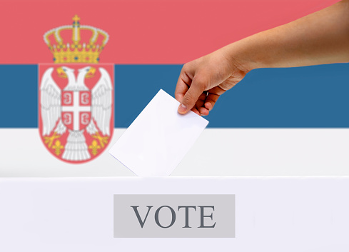 Male hand putting voting ballot into the vote box on blurred Serbian flag in background