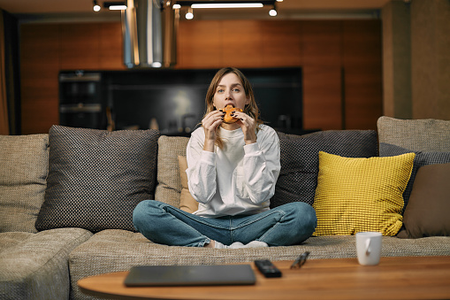 Young woman eating burger junk food watching tv series or movie, sitting on sofa in living room, resting at home. High quality photo