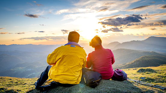 Back view of people in vibrant colour sportswear sitting above valley and enjoying panoramic view against sunset. Hiking in mountains and active lifestyle concept..