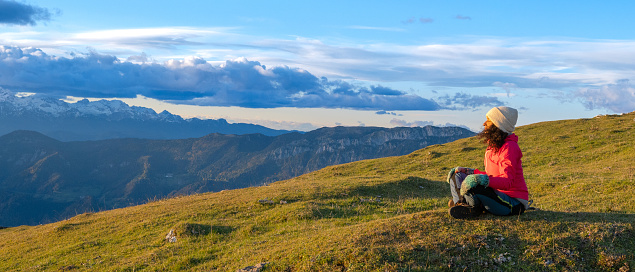 Side view of curly haired brunette sitting on grass and meditating in landscape illuminated by setting sun. Hiking in mountains and active lifestyle concept..