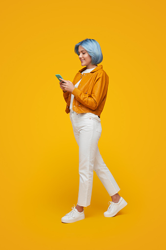 Side view of stylish female hipster with blue hair messaging on social media on mobile phone on yellow background in studio