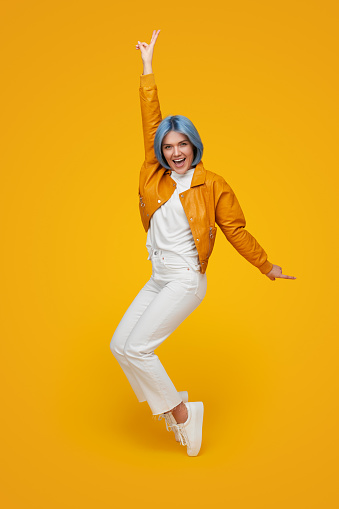 Side view full body of cheerful female hipster with blue hair and in trendy clothes, balancing on tiptoes while looking at camera on yellow background
