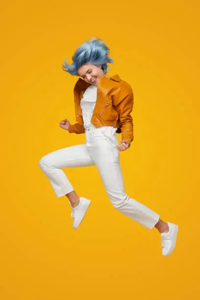 Full body of stylish teen female model with dyed hair wearing trendy leather jacket and white pants jumping against yellow background