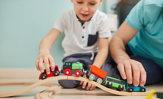 Crop little boy in casual clothes playing with toy train with wooden rails while sitting on floor with anonymous dad at home
