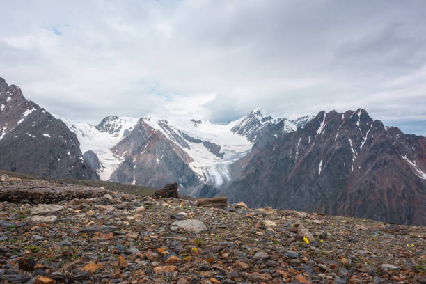 istock Dramatic landscape with high snowy mountain range with long glacier tongue and pointed peak in sunlight under cloudy sky. Atmospheric view from stone hill to large snow mountains at changeable weather 1375439963