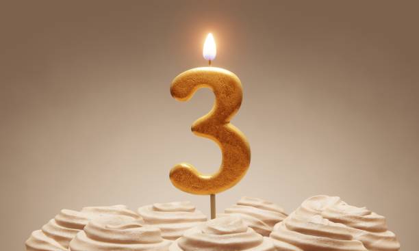 istock 3rd birthday or anniversary celebration. Lit golden number candle on cake with icing in neutral tones. 3D rendering 1375485894