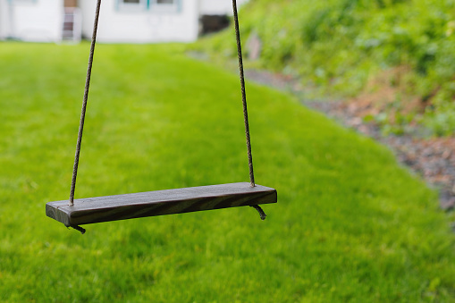 Empty wooden board rope-swing, hanging above green lawn and in front of white frame house.  Selective focus on swing.