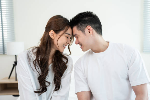 Portrait of Asian young couple sitting on bed and look at each other. Attractive beautiful new marriage man and woman in pajamas enjoy morning activity in bedroom at home. Family relationship concept. stock photo