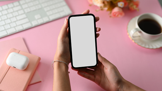 Close-up, Female hand holding phone white screen mockup over feminine pink office workspace table.