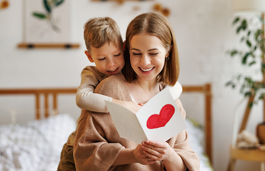 Cute little boy son congratulating his mom happy  woman with Mothers day, giving her handmade greeting postcard with red heart while sitting together on bed at home. Family holidays concept
