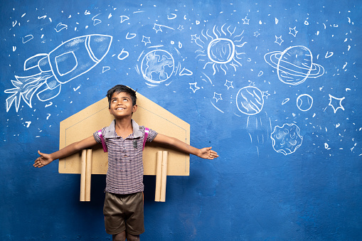 happy kid with cardboard rocket on back with space, universe and planets doodle drawing on wall - concept showing of childhood dream about astronaut or scientist.