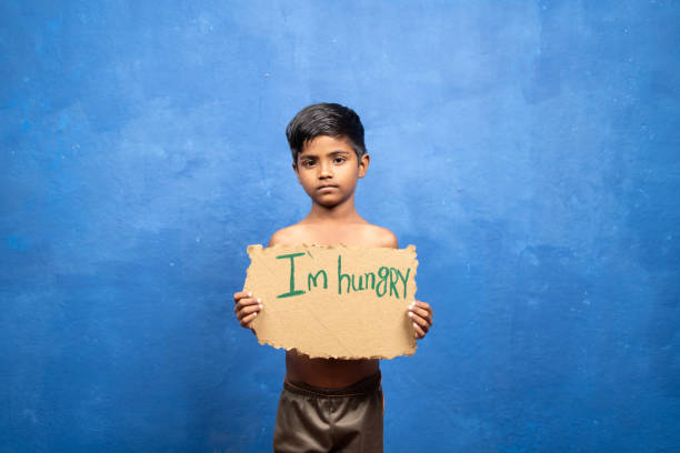 triste niño pobre indio sosteniendo tengo hambre letrero mirando la cámara en el fondo de blu con espacio de copia - concepto de pobreza y desnutrición. - malnourished fotografías e imágenes de stock