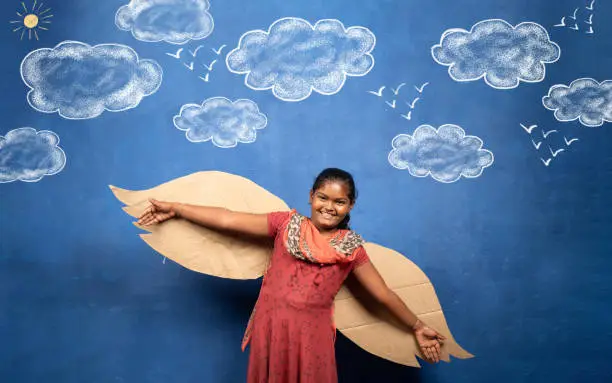 Photo of excited girl with wings flying on cloud doodle drawing on blue background - concept of childhood freedom, poverty and motivation