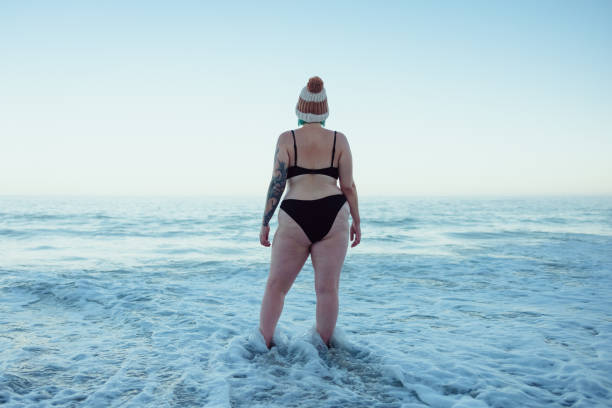 Female winter bather watching the sea waves at the beach Female winter bather watching the sea waves at the beach. Rearview of an unrecognisable winter bather wearing swimwear and a wooly hat. Woman standing in the cold sea water. woman cold water swimming stock pictures, royalty-free photos & images