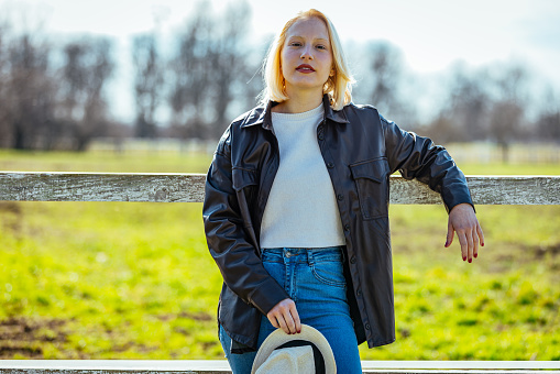 A woman with a cowboy hat, leaning against a village fence next to a horse pasture, looks confident.