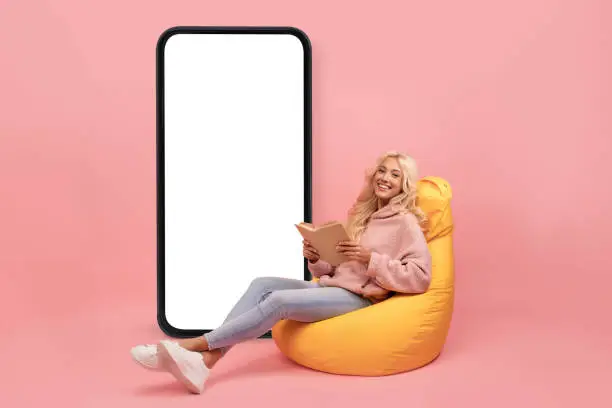 Photo of Online library or book store. Young lady reading textbook in bean bag chair, offering mockup on smartphone screen