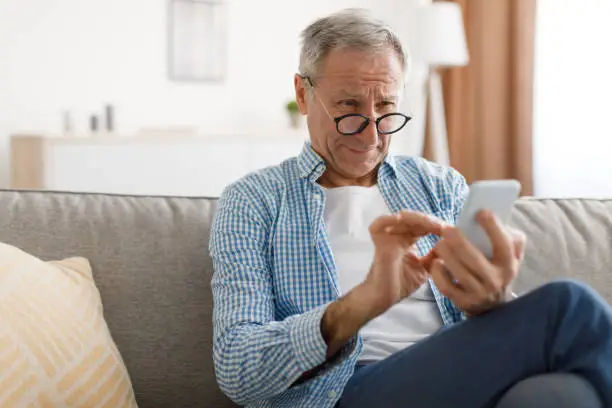 Poor Eyesight. Senior Man Squinting Eyes Reading Message On Phone Wearing Eyeglasses Having Problems With Vision Sitting On Couch. Ophtalmic Issue, Bad Sight In Older Age, Macular Degeneration Concept
