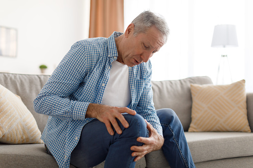 Osteoarthritis. Senior man holding touching his knee feeling acute pain, sick mature male suffering from aching leg sitting on couch at home. Old age health problem concept. Rheumatoid arthritis