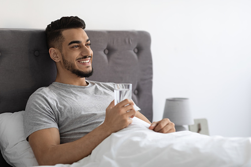 Happy Handsome Arab Man Sitting In Bed And Holding Glass With Water, Smiling Young Middle Eastern Guy Resting In Bedroom, Enjoying Healthy Drink After Waking Up In The Morning, Closeup, Copy Space